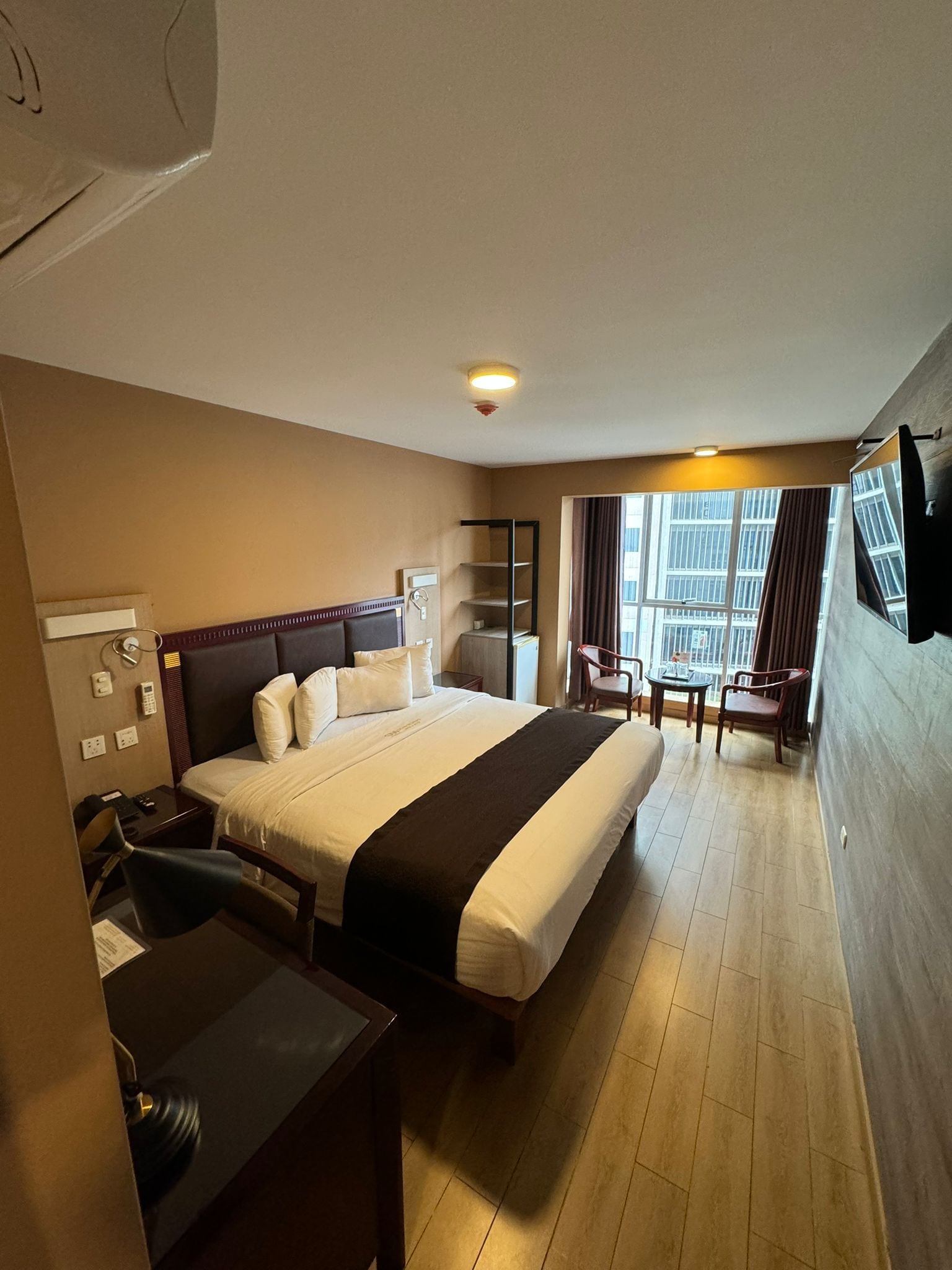 A hotel room with a large bed, bedside tables, a desk, a chair, a small table with chairs, a TV, and a window with curtains.