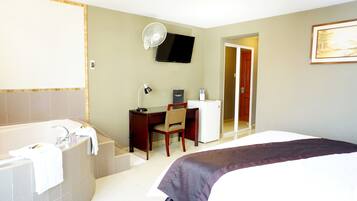 A hotel room with a large bed, a desk with a chair, a small refrigerator, a television mounted on the wall, and a bathroom with a bathtub and a mirror.