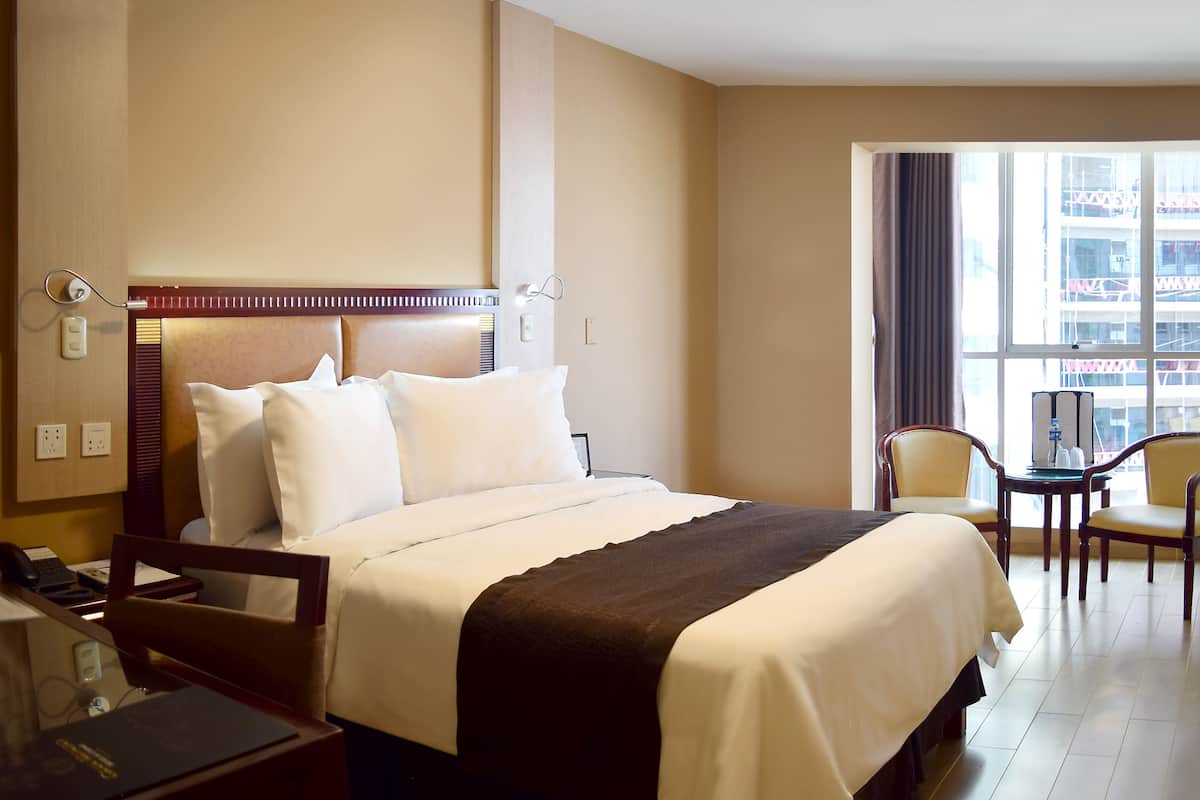 Habitación de hotel con cama, mesita de noche, lámpara y vista a la ciudad por la ventana.
