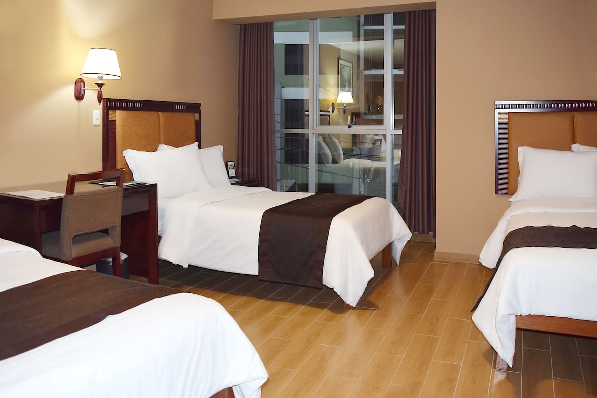 Habitación de hotel con tres camas, un escritorio, una silla y un ventanal con cortinas.