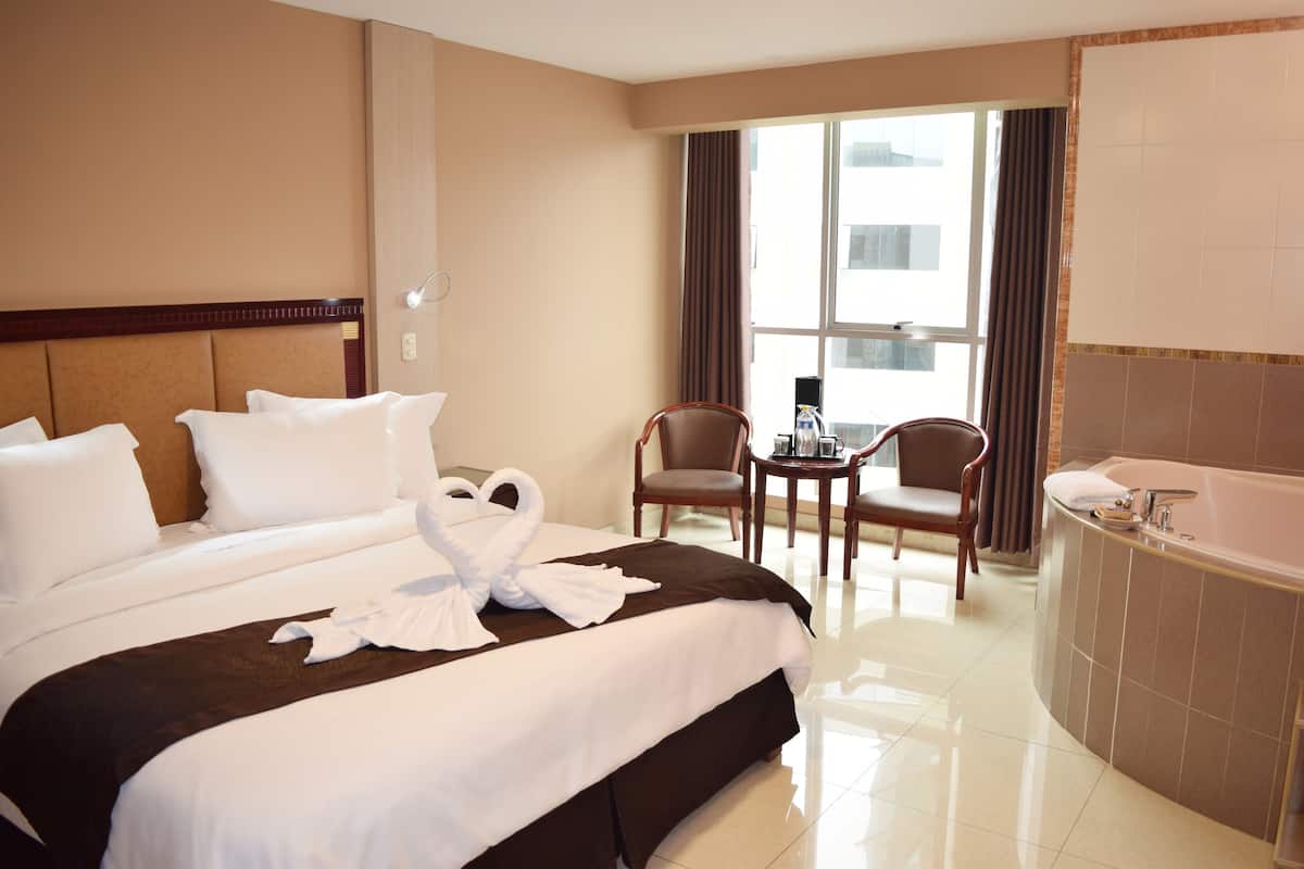 Habitación de hotel con una cama grande, un espacio de descanso con dos sillones, una mesita y un baño independiente con bañera.