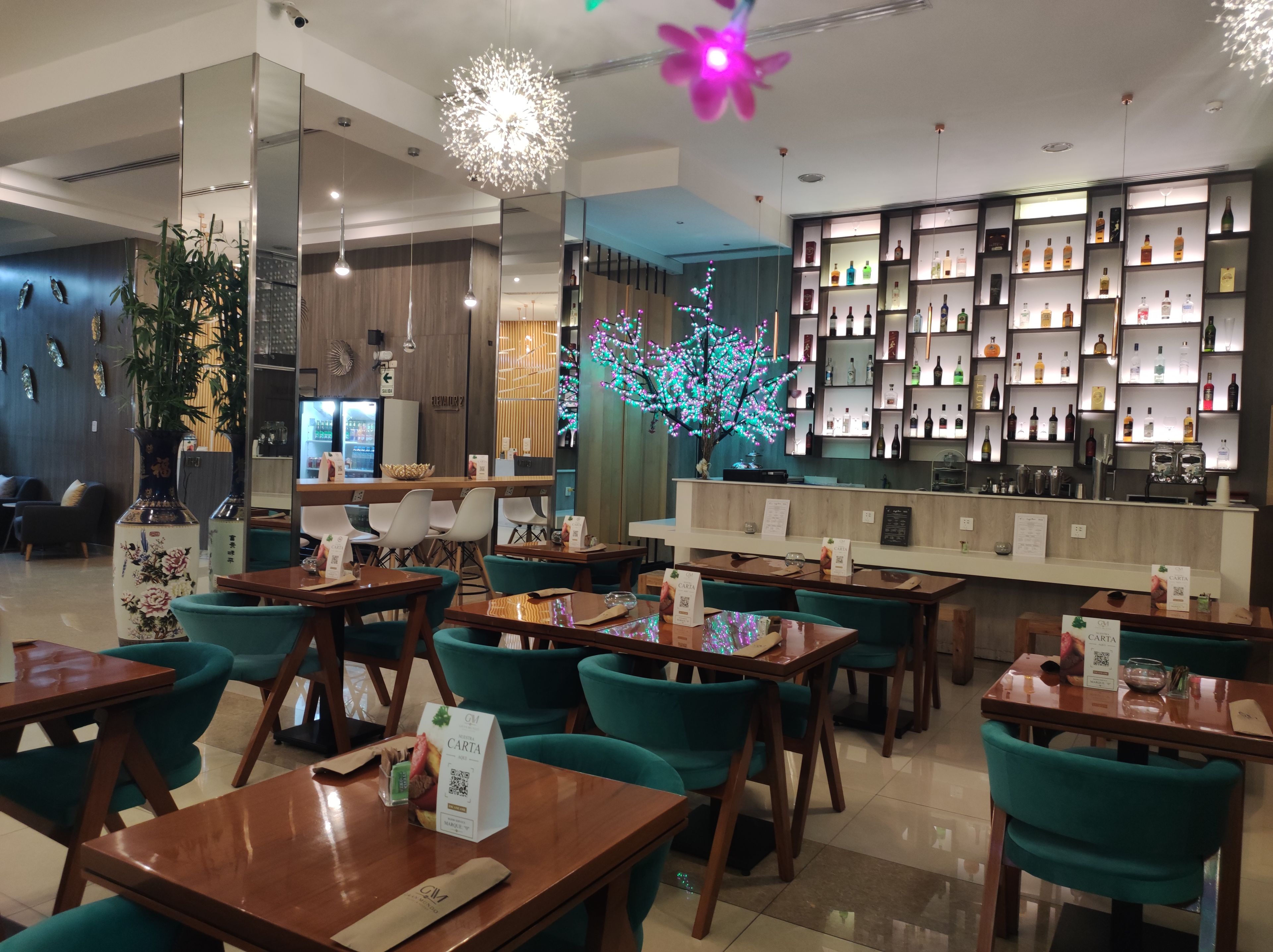 Ein Restaurant mit Holztischen und türkisfarbenen Stühlen, ein Barbereich mit Regalen voller Flaschen und dekorativer Beleuchtung.
