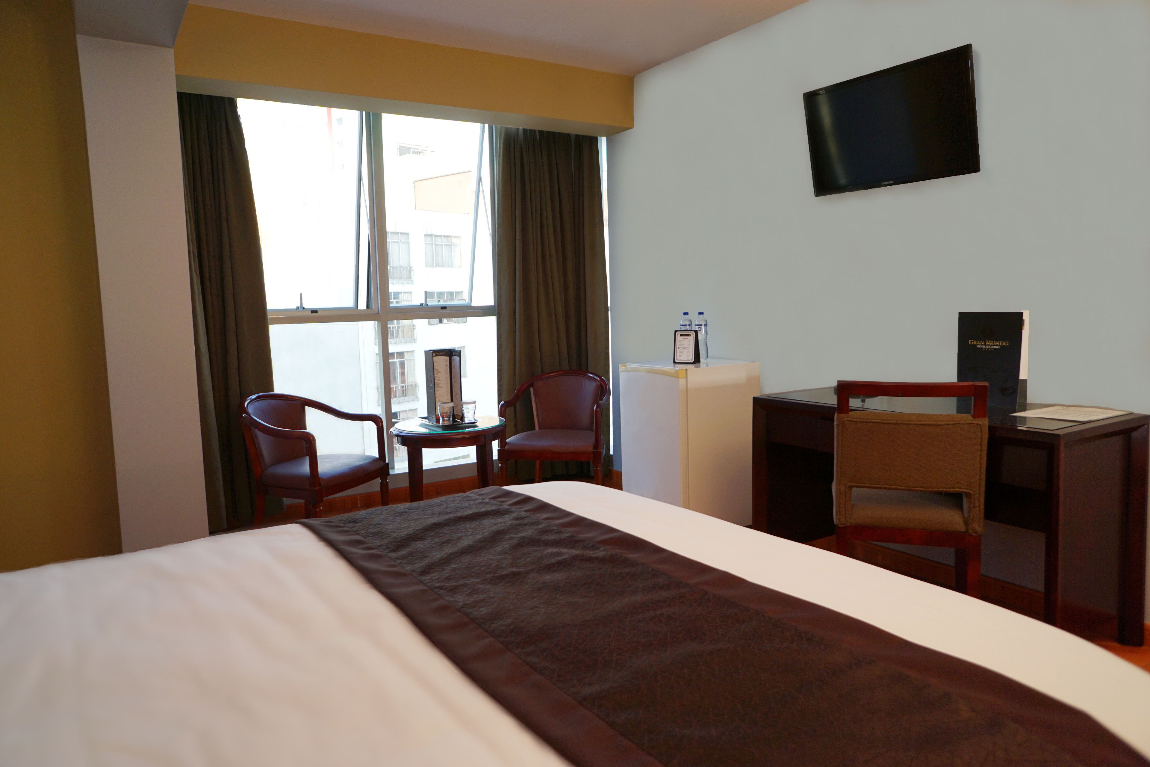 A hotel room with a bed, two chairs, a desk, a television, and a window with curtains.