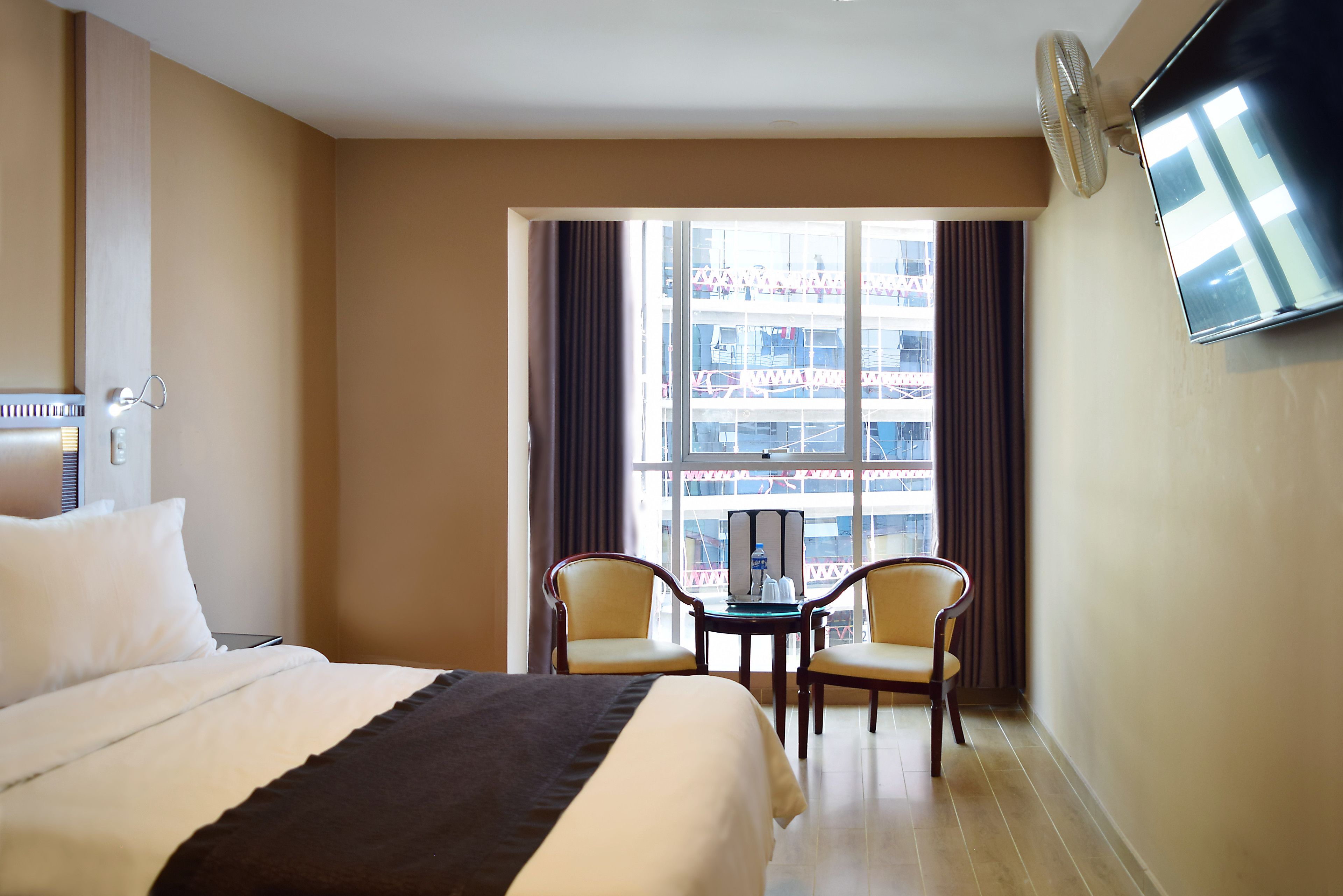 Habitación de hotel con cama, dos sillas, una mesa con una botella y televisión en la pared.