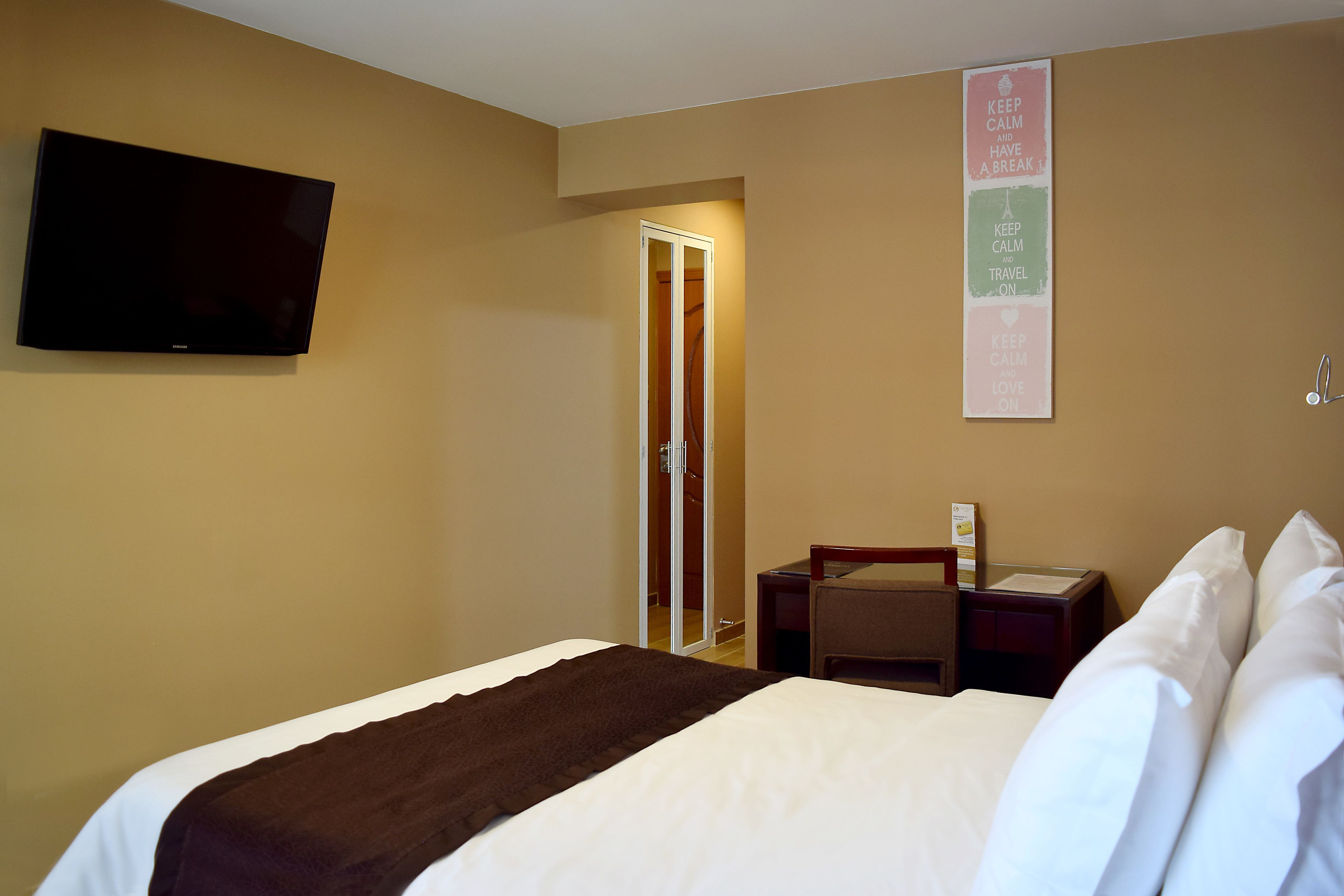 A hotel room with a bed, a flat-screen TV mounted on the wall, a nightstand, a desk, and a door leading to a bathroom.