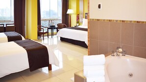 Habitación de hotel con dos camas, una zona de estar con una silla y una mesa, una bañera grande y vistas a la ciudad a través de las ventanas.