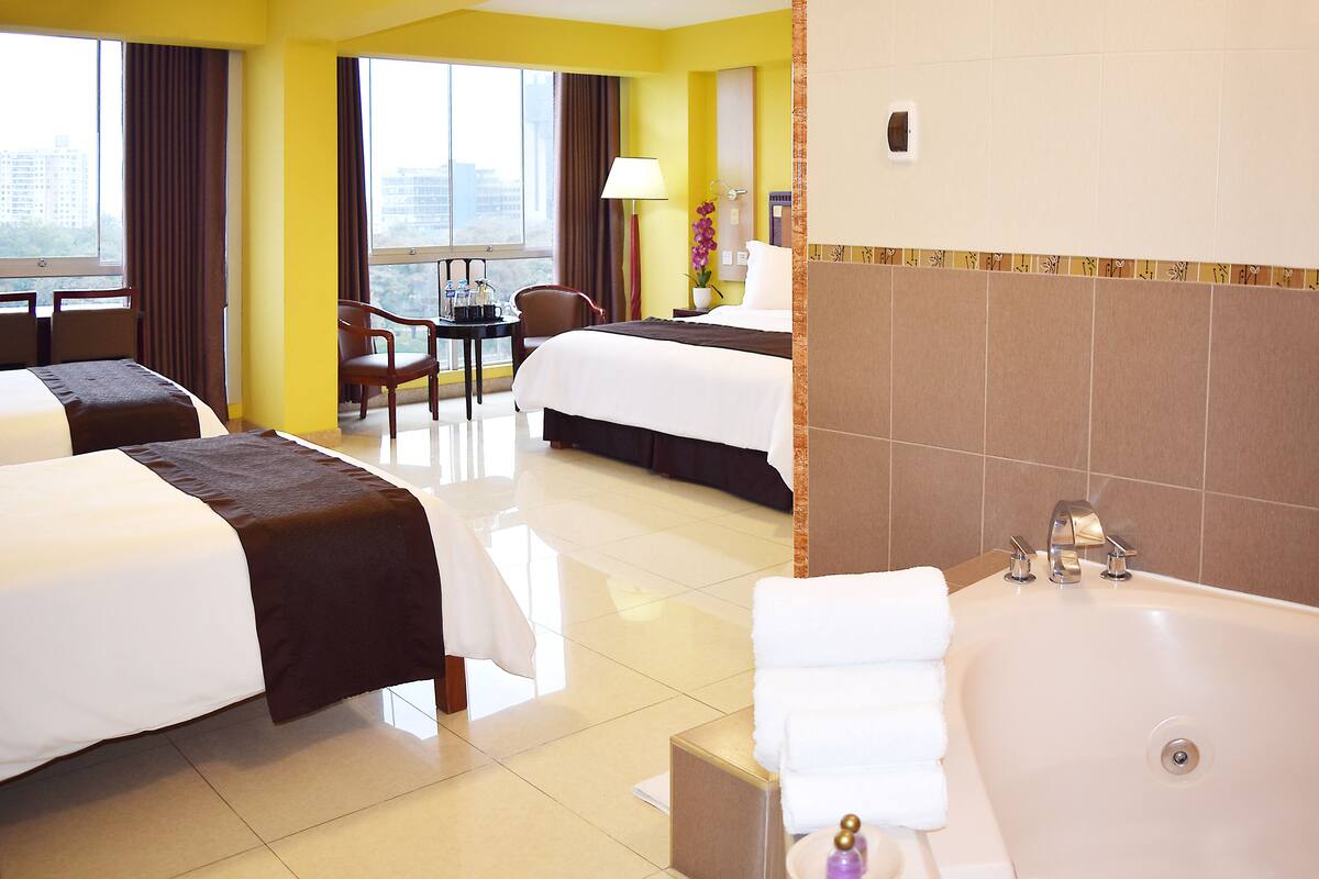 Habitación de hotel con dos camas, una zona de estar con una silla y una mesa, una bañera grande y vistas a la ciudad a través de las ventanas.
