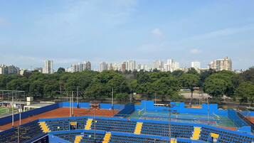Un court de tennis avec des sièges bleus et jaunes, entouré d’arbres et d’immeubles de la ville.