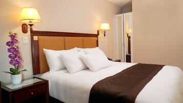 Habitación de hotel con una cama grande, dos lámparas de noche, una mesita de noche con un florero y una puerta a la derecha.
