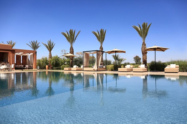 Un área junto a la piscina con sillones de descanso y sombrillas, rodeada de palmeras.