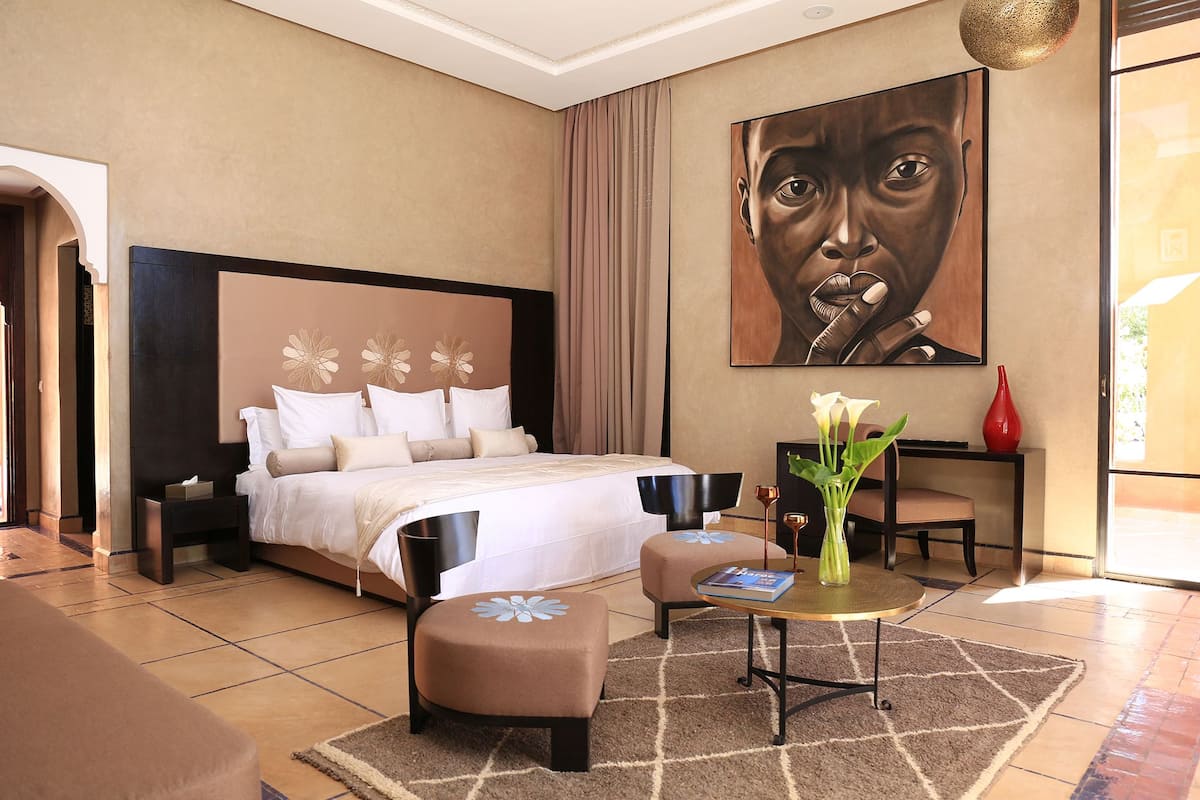 A hotel room with a large bed, a sitting area with a table and chairs, a painting on the wall, and a vase with flowers on a side table.