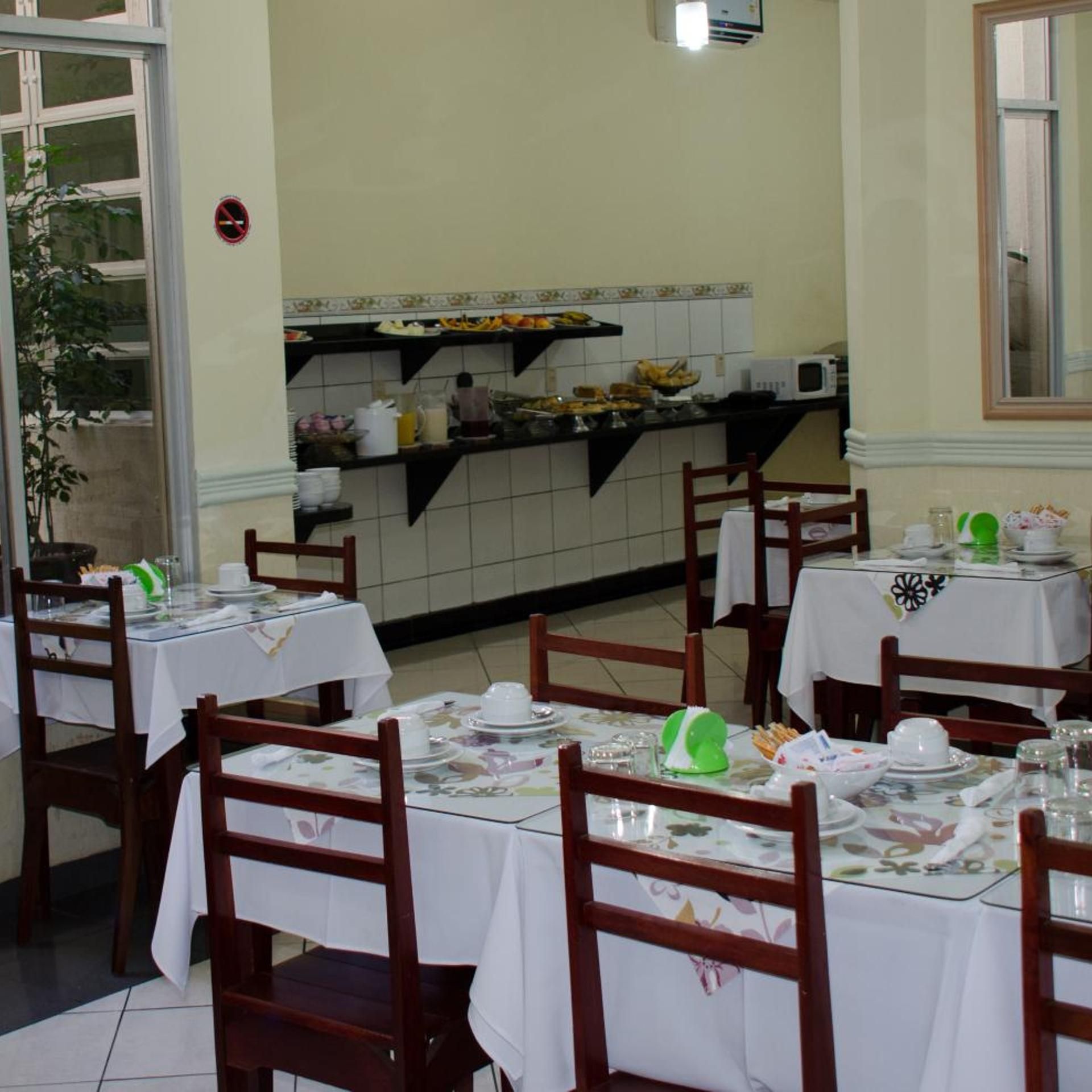 Uma área de refeições com mesas preparadas para o serviço, uma cozinha ao fundo e uma planta perto da entrada.