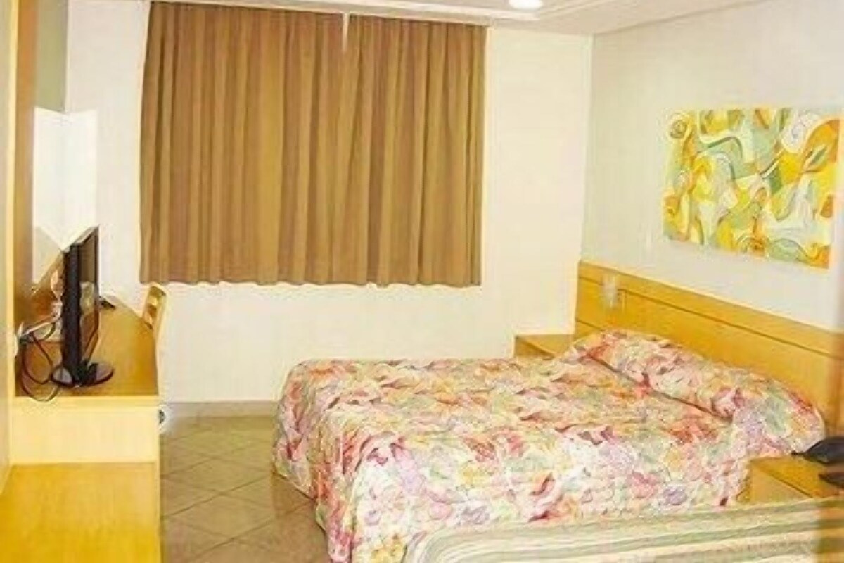 Une chambre d’hôtel avec un lit, une télévision, une fenêtre avec des rideaux et un tableau au mur.