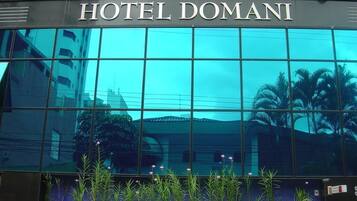 Exterior del Hotel Domani, con un amplio ventanal de vidrio que refleja los edificios y las palmeras circundantes.