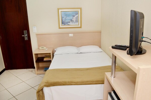 A hotel room with a bed, a bedside table with a phone, a framed picture on the wall, and a desk with a television and remote controls. - Steinhausen Hotel (Blumenau)