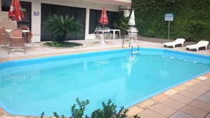 Une piscine entourée de chaises longues et de parasols, le tout bordé d’un bâtiment doté d’une terrasse carrelée et d’une haie.