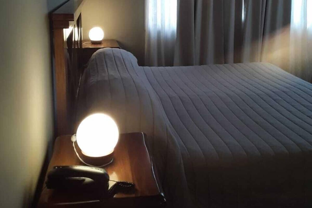 Ein Hotelzimmer mit Bett, Nachttisch mit Lampe, Telefon und Vorhängen an den Fenstern.