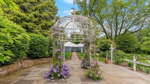 Ein gepflasterter Weg führt zu einem weißen Gazebo mit violetten Blumen, umgeben von üppigem Grün.