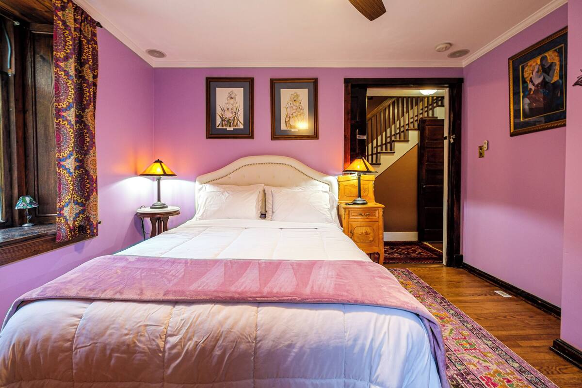 Une chambre à coucher avec un grand lit, deux lampes de chevet, une fenêtre avec des rideaux et un escalier menant à une autre pièce.