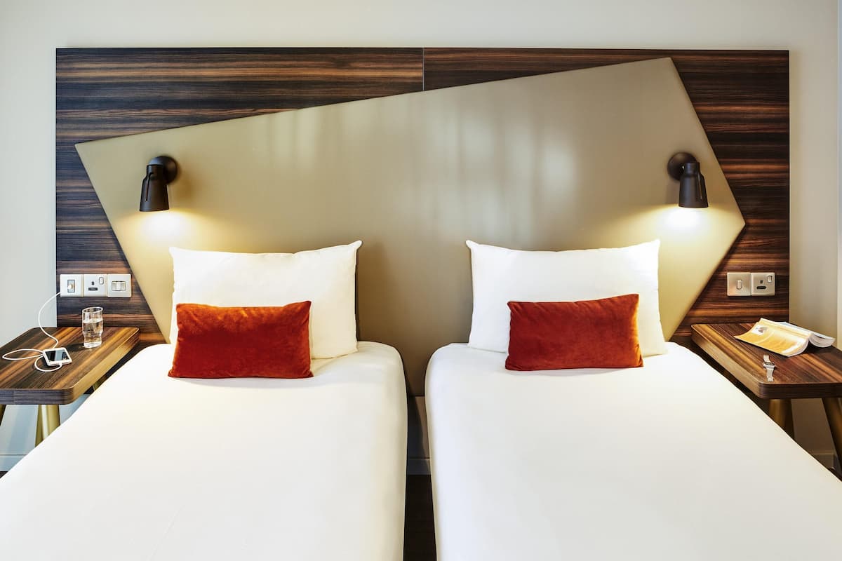 Habitación de hotel con dos camas, cada una con una almohada y una mesita de noche con lámpara.