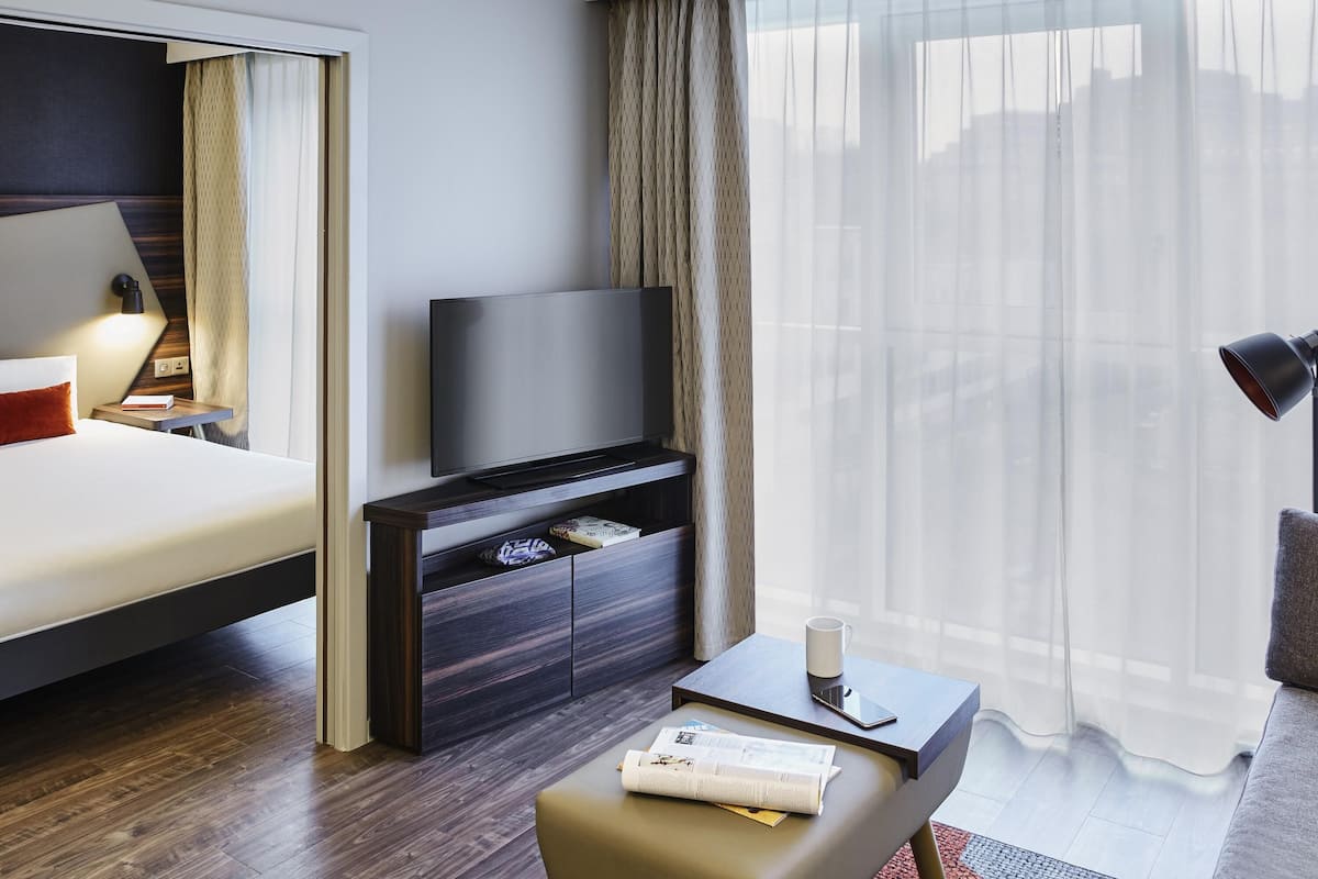 Una habitación de hotel moderna con una cama, un televisor sobre un soporte de madera, una mesita con una taza y un teléfono inteligente, y una ventana con cortinas translúcidas.