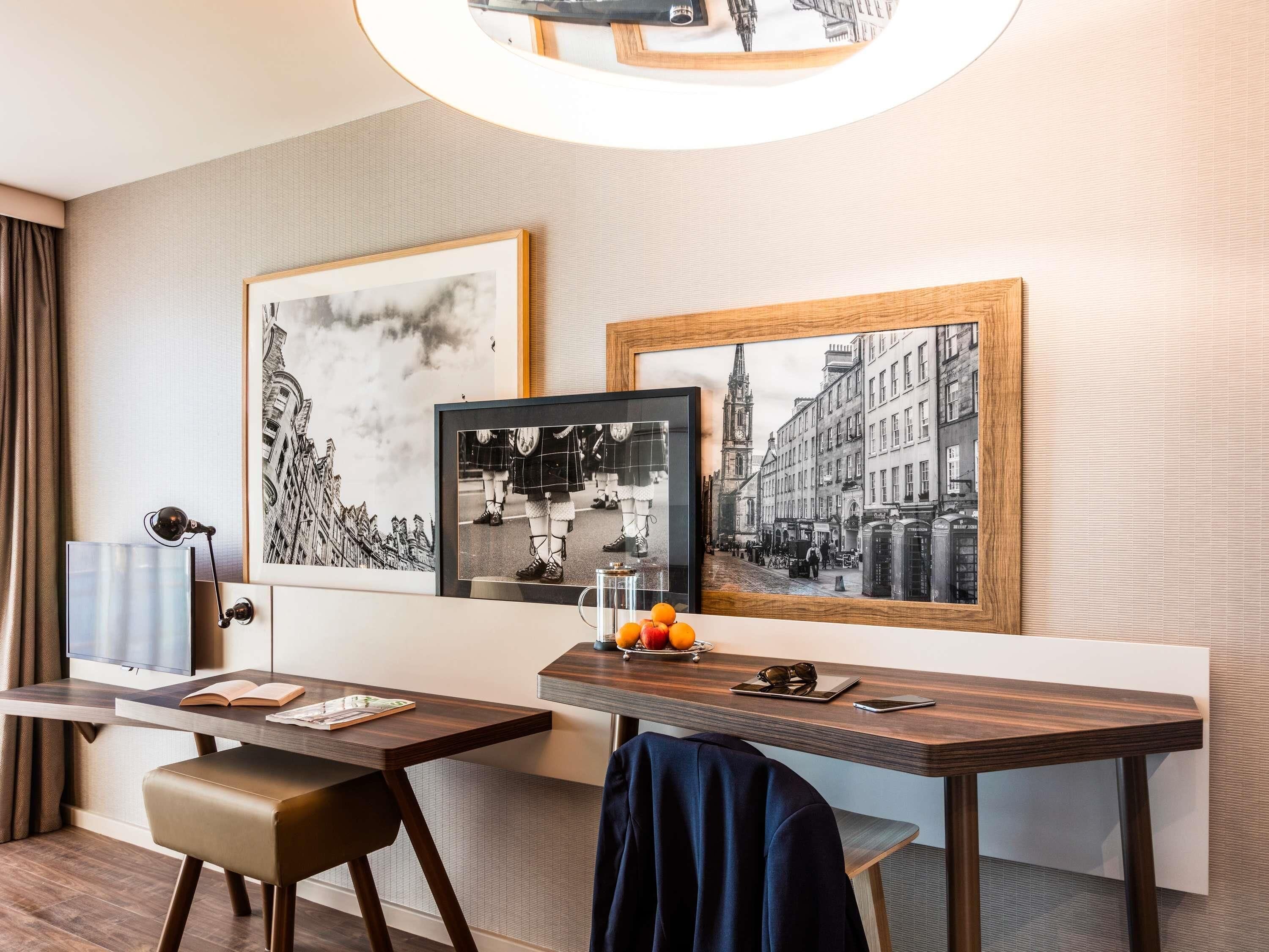 A hotel room with a desk, a chair, a bed, a bedside table with a lamp, and two framed pictures on the wall.