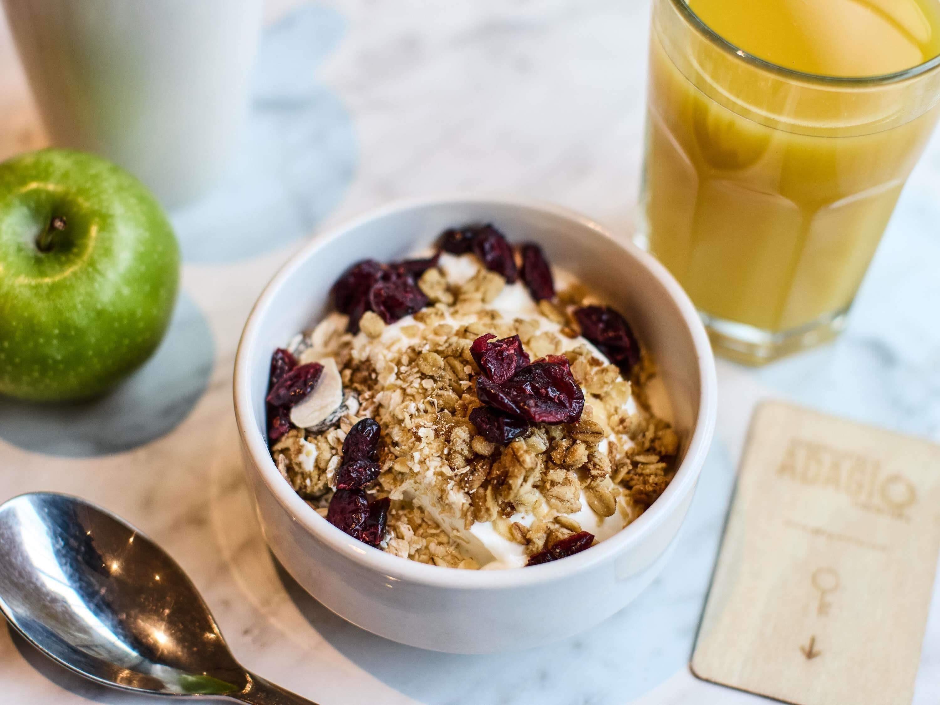 A bowl of granola with dried fruit and nuts, a glass of orange juice, a green apple, and a spoon on a marble surface.