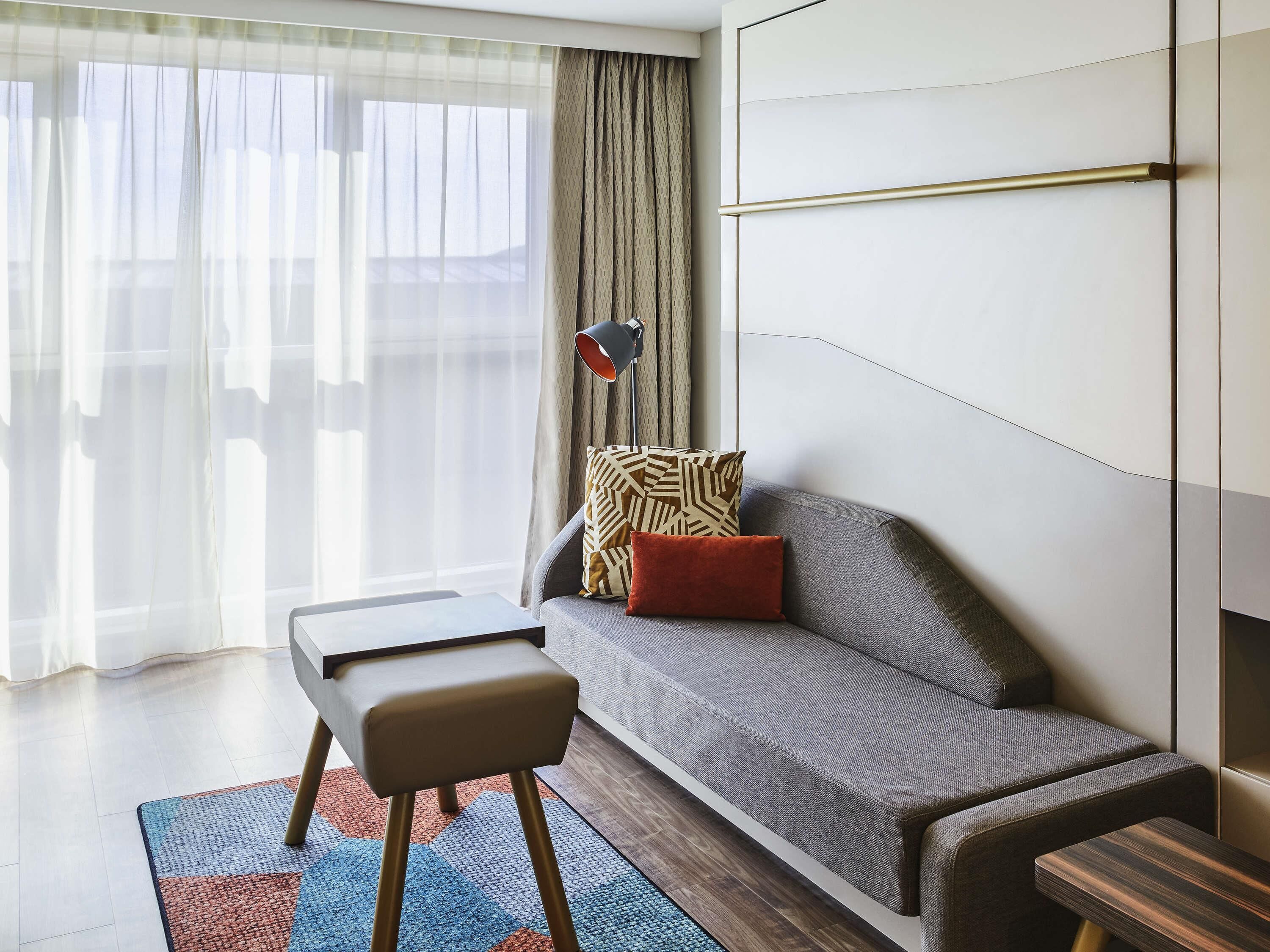 A modern living room with a grey sofa, a small wooden bench, a patterned throw pillow, and a floor lamp.