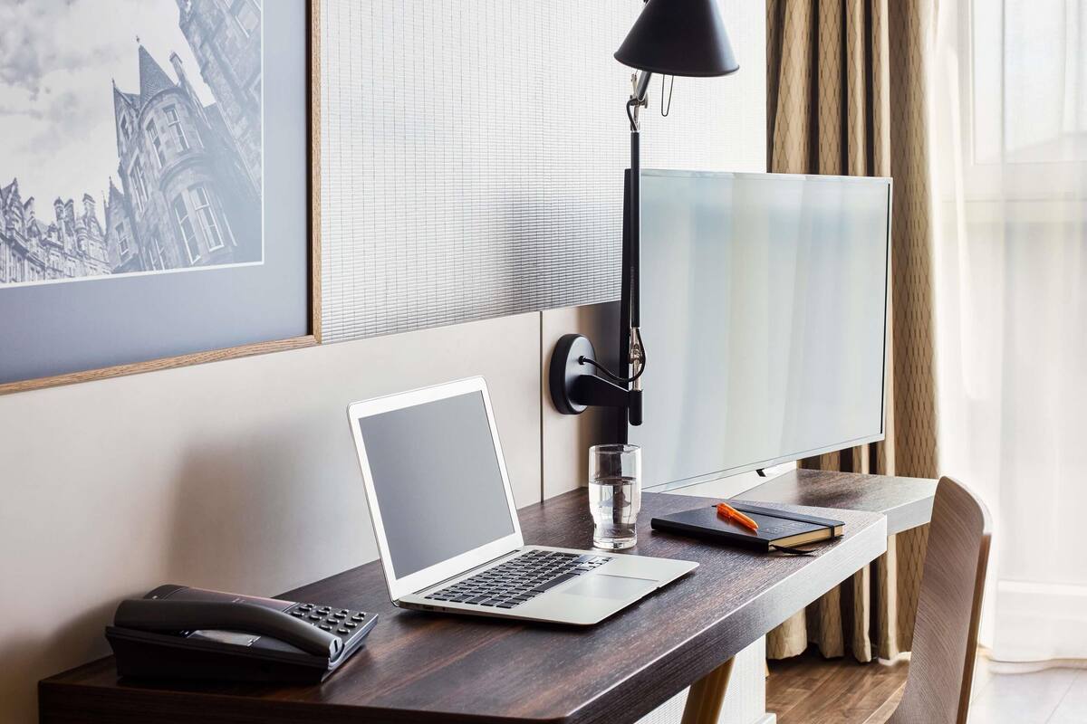 Habitación de hotel con un escritorio que tiene una computadora portátil, un teléfono, un vaso de agua, una libreta y un bolígrafo. Una lámpara de escritorio negra está sobre el escritorio. La habitación tiene un ventanal grande con cortinas, y un cuadro enmarcado en la pared.