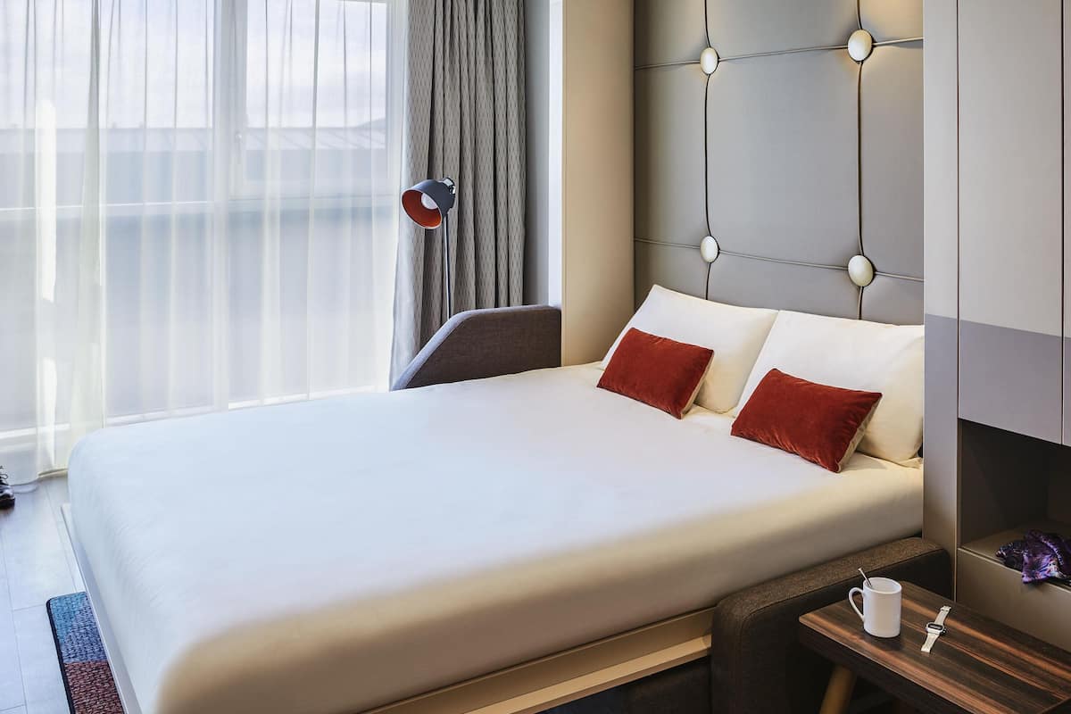 A hotel room with a large bed, a bedside table with a cup and a watch, a lamp, and a view of the outside through sheer curtains.
