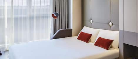 Quarto de hotel com uma cama grande, uma mesa de cabeceira com uma chávena e um relógio, uma lâmpada e vista para o exterior através de cortinas translúcidas.