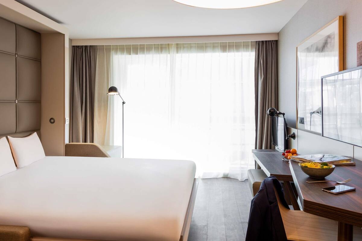Habitación de hotel con una cama grande, un escritorio con un frutero, una lámpara y un ventanal con cortinas.