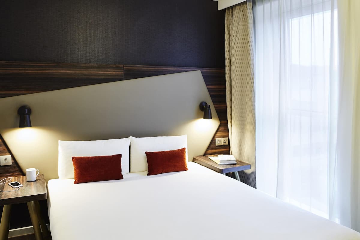 Habitación de hotel con una cama grande, dos lámparas de noche, una mesita de noche y una ventana con cortinas.
