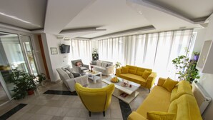 A modern living room with a yellow sofa, a grey sofa, and a yellow armchair. There is a coffee table in the center with a fruit bowl on it. The room has large windows with sheer curtains and a flat-screen TV mounted on the wall.