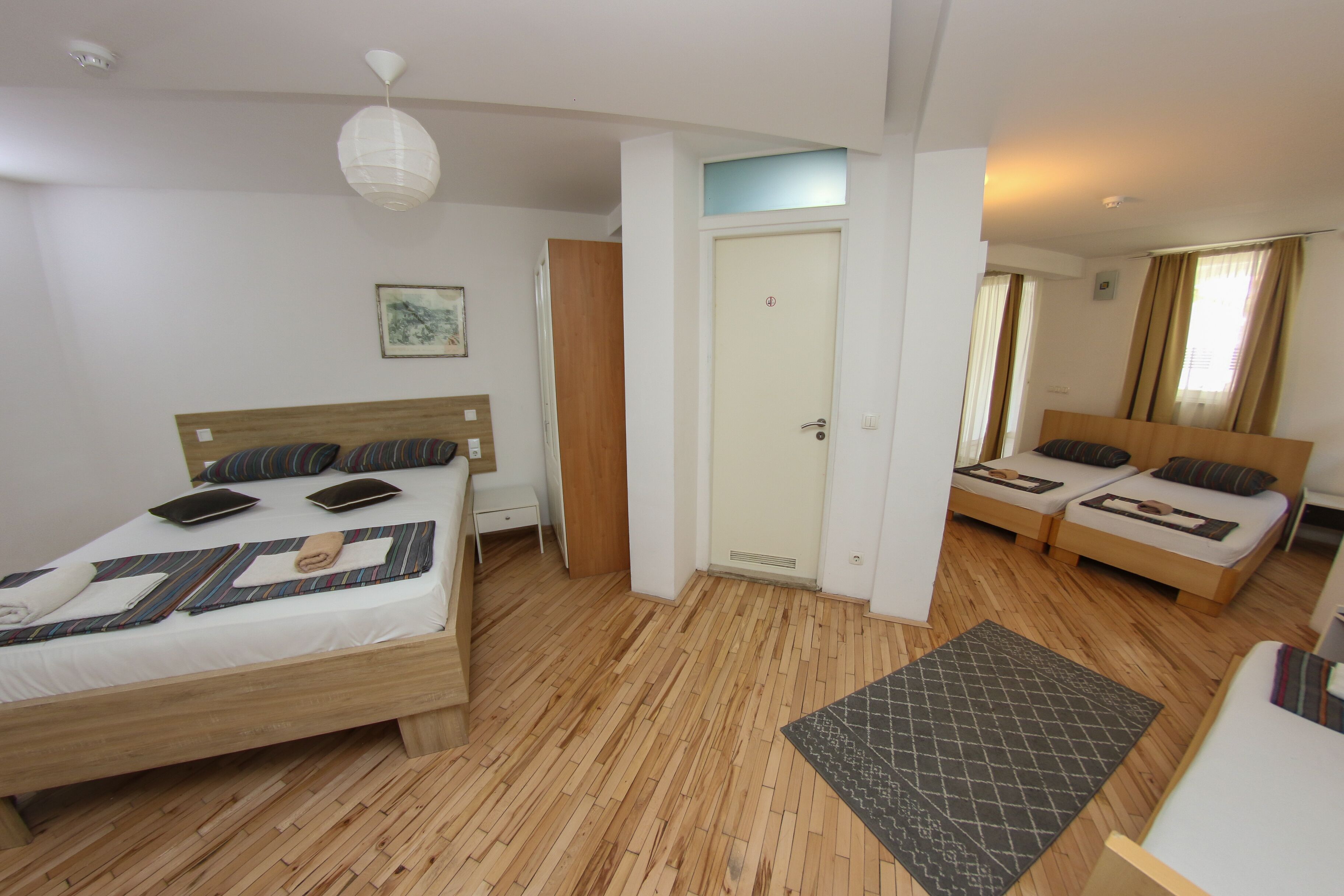 Une chambre d’hôtel avec un plancher en bois, un lit avec une tête de lit, une table de chevet, une petite table, une lampe fixée au mur et une porte donnant sur une autre pièce.