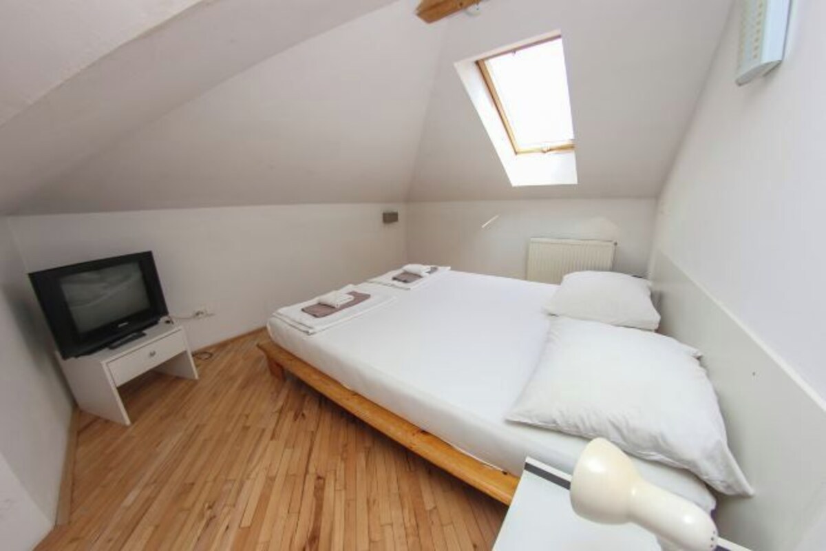 A small, single-room apartment with a sloped ceiling, a bed with white bedding, a small bedside table with a television, and a window providing natural light.