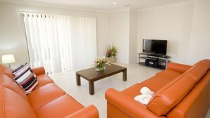 A living room with orange leather sofas, a wooden coffee table, a flat-screen TV on a stand, and a window with white curtains. - Stoneyfell (POINT COOK)