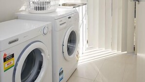 A laundry area with two washing machines and a basket, a tiled floor, and a door with vertical blinds. - Stoneyfell (POINT COOK)