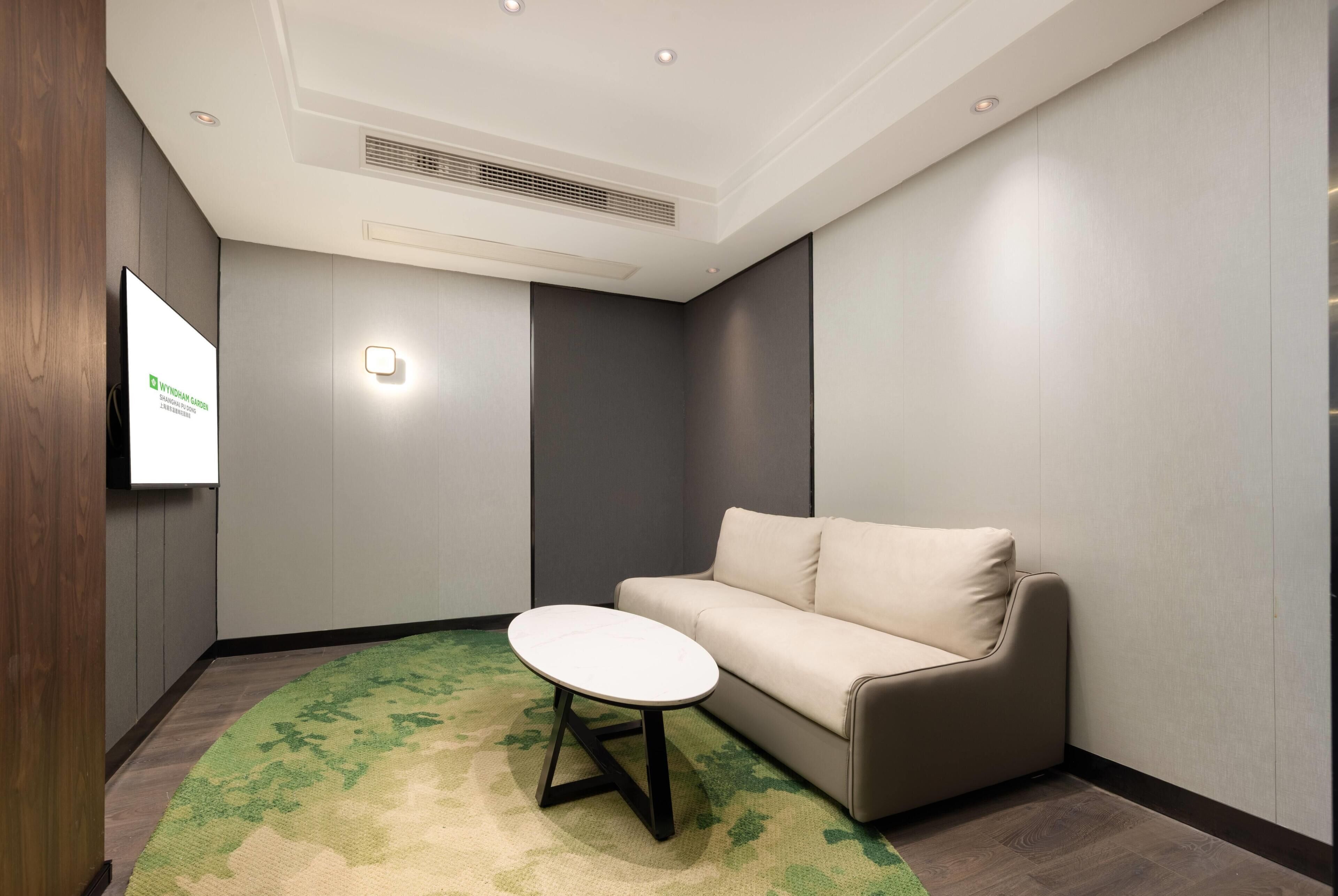 A conference room with a white table, a beige sofa, a green patterned carpet, and a mounted screen.