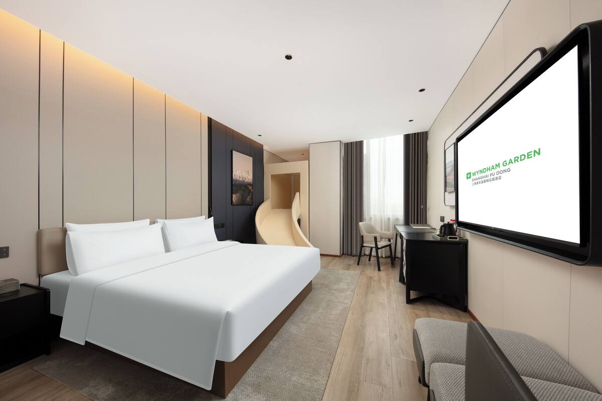 Une chambre d’hôtel moderne équipée d’un grand lit, d’un bureau, d’une chaise et d’un écran de télévision affichant le texte « WYNHAM GARDEN SHANGHAI PU DONG ».