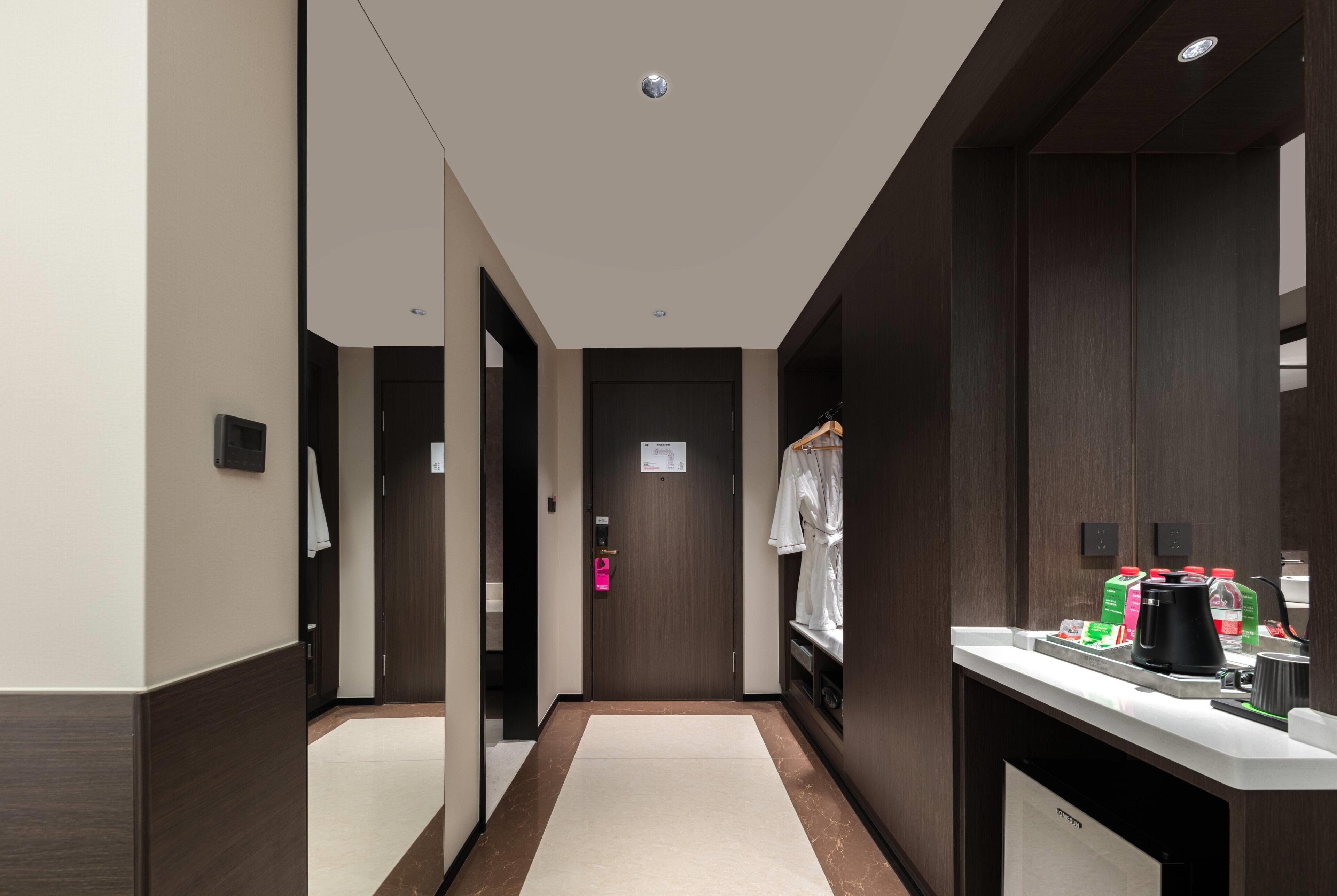 A hotel corridor with a wooden floor, a white wall, a door with a pink lock, and a counter with a coffee maker and a water dispenser.