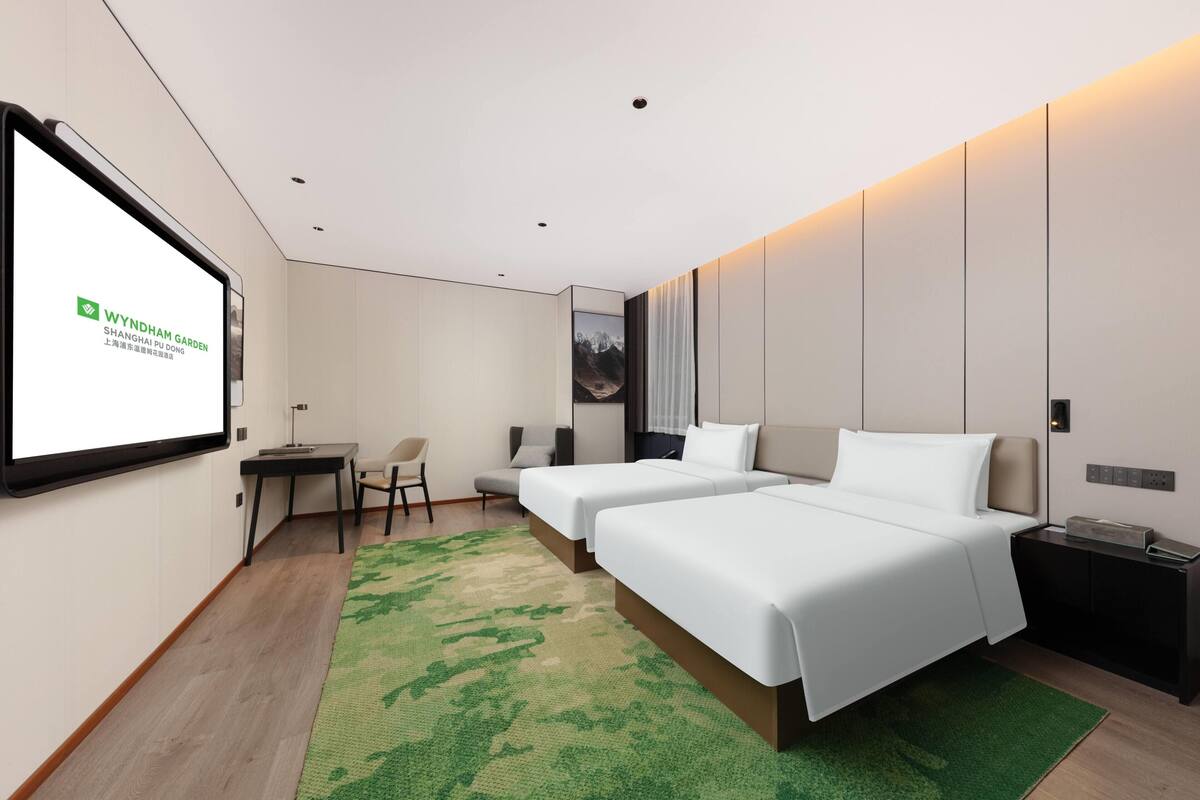 Une chambre d’hôtel moderne équipée de deux lits, d’un bureau, d’une chaise et d’un grand écran affichant le logo WYNDHAM GARDEN.
