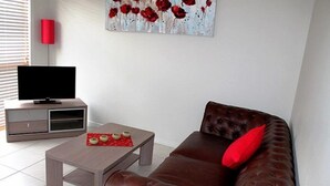 A living room with a brown leather sofa, a wooden coffee table, a flat-screen TV on a stand, and a red vase on the wall. - City Cottage (GOWANBRAE)