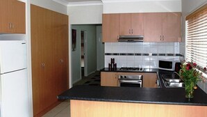 A modern kitchen with wooden cabinets, a black countertop, stainless steel appliances, and a tiled backsplash. - City Cottage (GOWANBRAE)