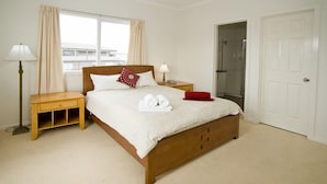 A neatly arranged bedroom with a wooden bed, two bedside tables, a window with curtains, a door, and a visible bathroom. - City Condo (GOWANBRAE)