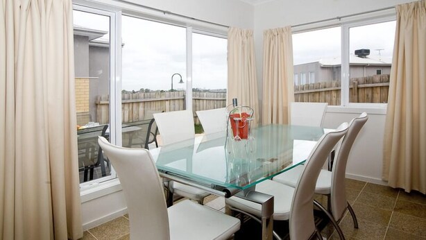 A dining area with a glass table and white chairs, large windows with curtains, and a view of a wooden fence. - City Condo (GOWANBRAE)