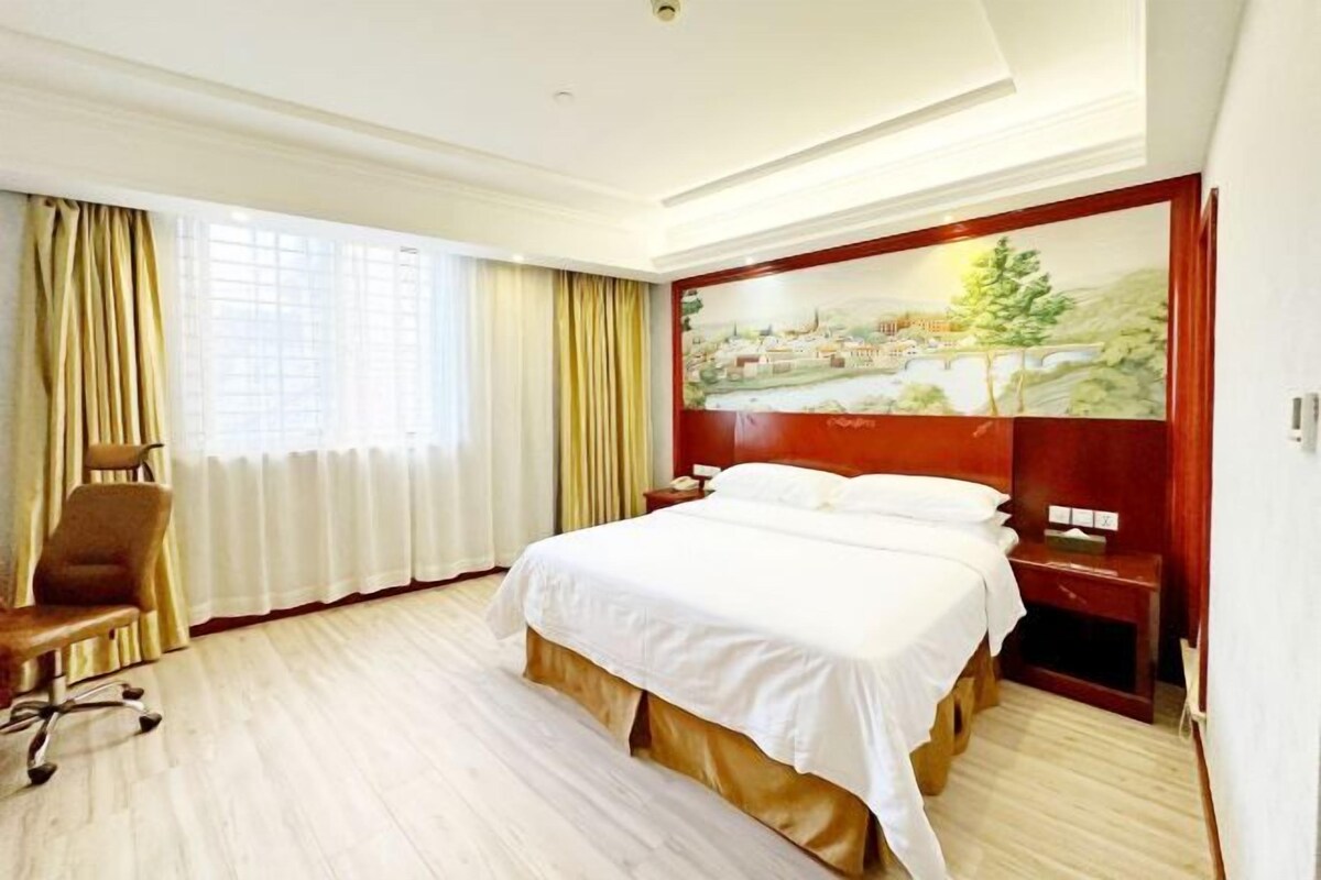 A hotel room with a bed, a chair, a nightstand, a painting on the wall, and a window with curtains.