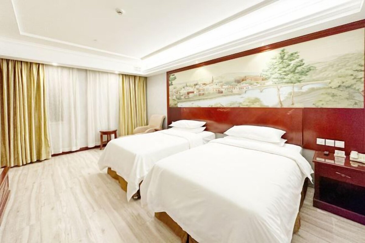 A hotel room with two beds, a painting on the wall, a nightstand, and a window with curtains.