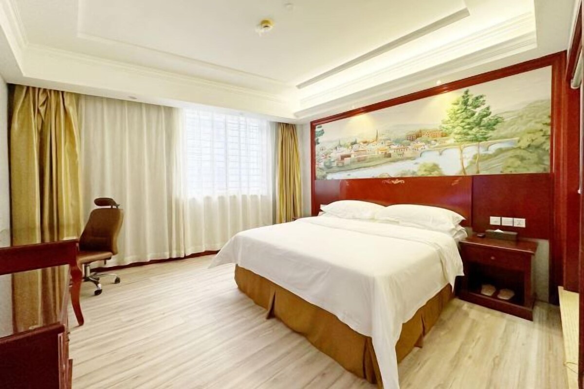 A hotel room with a large bed, a desk, a chair, and a painting on the wall.