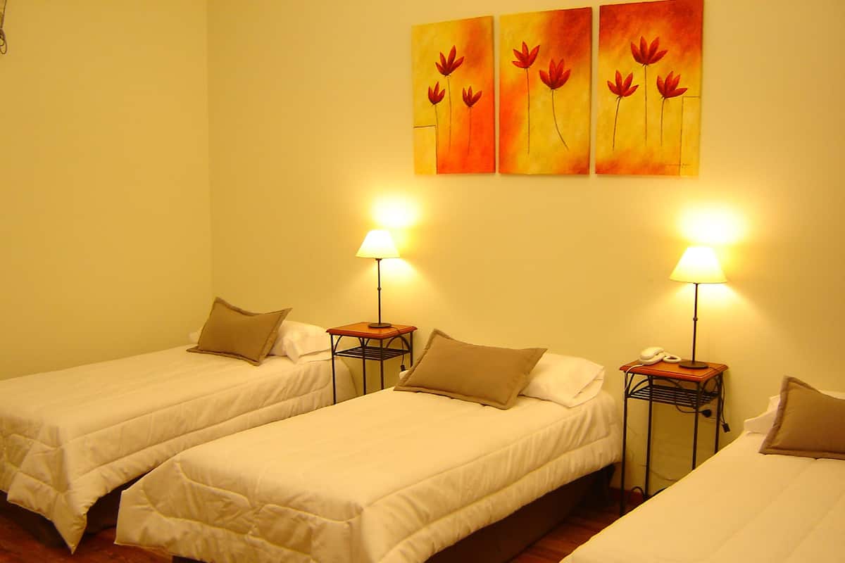 A hotel room with three single beds, each with a bedside table and lamp.